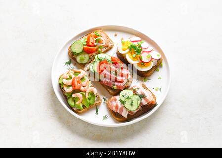 Verschiedene Sandwiches mit Fleisch, Gemüse, Meeresfrüchten. Auswahl offener Sandwiches auf Platte über hellem Steingrund mit freiem Textbereich. Schmackhaftes Heils Stockfoto