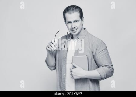 Ein Mann in einem Hemd auf gelbem Hintergrund hält ein Buch, während er seine Brille abnimmt und schaut auf die Kamera. Schwarz und Weiß. Nahaufnahme. Stockfoto