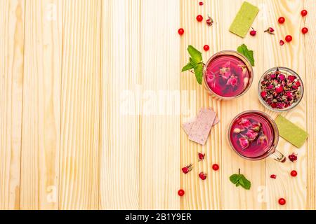 Im Sommer köstlicher grüner Rosen-Tee. Heißes romantisches Getränk in der Tasse mit Himbeer-Schokolade, rosafarbenen Kronblättern und Knospen, roten Beeren viburnum, Minze. Holzplatten B. Stockfoto