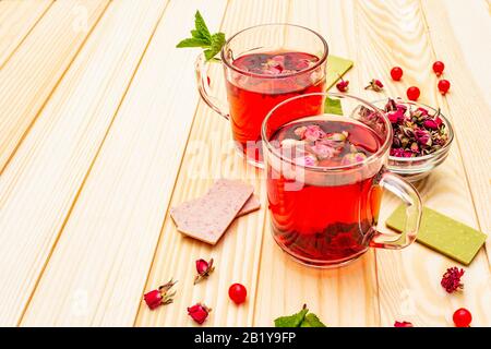 Im Sommer köstlicher grüner Rosen-Tee. Heißes romantisches Getränk in der Tasse mit Himbeer-Schokolade, rosafarbenen Kronblättern und Knospen, roten Beeren viburnum, Minze. Holzplatten B. Stockfoto