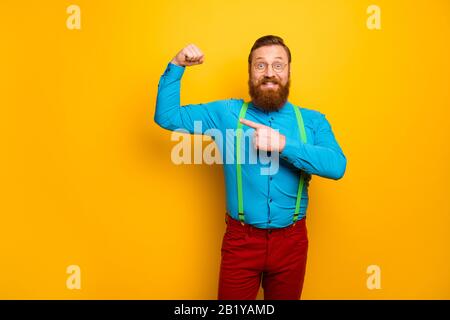 Sehen Sie. Foto von attraktivem lustigen Toothy Kerl Heben Sie den Faustarm an und zeigen den Finger an, der perfekte Form zeigt Bizeps tragen blaues Hemd grüne Hosenträger rote Hosen Stockfoto