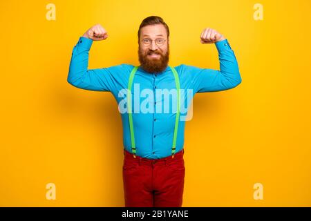 Foto von attraktivem lustigen Toothy Kerl hebt Fäuste Arme mit perfekter Form Bizeps nach dem Training Sporthalle tragen Sie blaues Hemd grüne Hosenträger rote Hosen Stockfoto