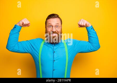 Foto von attraktivem Toothy Kerl hebt Fäuste Arme mit erstaunlicher Form große Bizeps nach dem Training Gym tragen blaue Hemd grüne Hosenträger isoliert Stockfoto