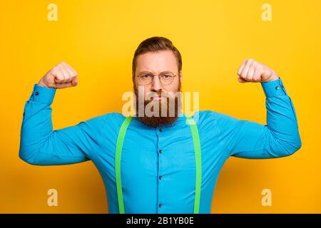 Foto des attraktiven lustigen Kerls hebt Fäuste schwer Arme mit perfekter Form starke Bizeps Sport süchtig tragen blaues Hemd grüne Hosenträger Stockfoto