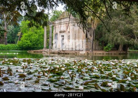 Das Gebäude wurde inmitten eines Teiches mit Lilienstätten im Englischen Garten des Königlichen Palastes von Caserta, Italien, abgebrochen. Аn Ruinen eines Tempels. Stockfoto