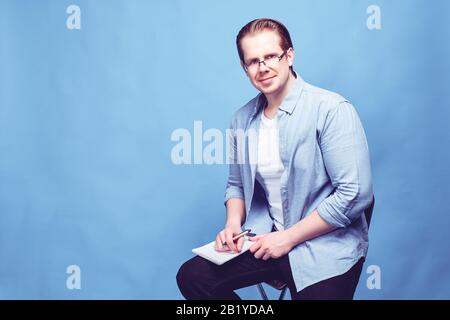 Ein Mann in legerer Kleidung mit Brille auf blauem Hintergrund sitzt auf einem Stuhl und sieht die Kamera an, während er ein Notizbuch hält. Nahaufnahme. Stockfoto