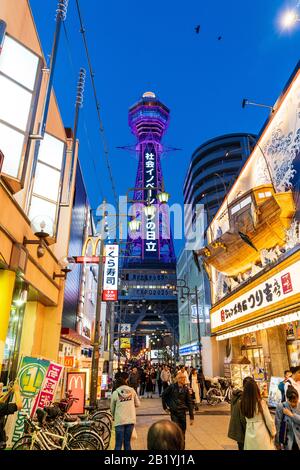 Die belebte Fußgängerzone vor dem Jumbo Fischerboot Tsurikichi-Restaurant mit dem Tsutenkaku Turm überragt. Nacht, Shinsekai in Osaka. Stockfoto