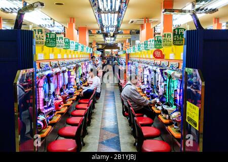 Innenansicht am Gang einer japanischen Pachinko-Stube mit zwei Reihen von Spielautomaten und einigen darauf spielenden Männern. Nachtzeit. Stockfoto