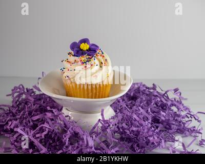 Vanillecupcake mit weißem Frost und einer violetten Blume und bestreut auf einem weißen Gericht, das von violettem Papier umgeben ist. Festlich, hübsch und lecker Stockfoto