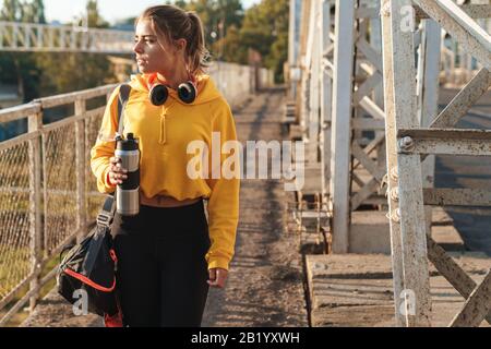 Wunderschönes junges Sportmädchen, das Sportswear auf einer Brücke im Freien trägt, Tasche trägt und Wasserflasche hält Stockfoto