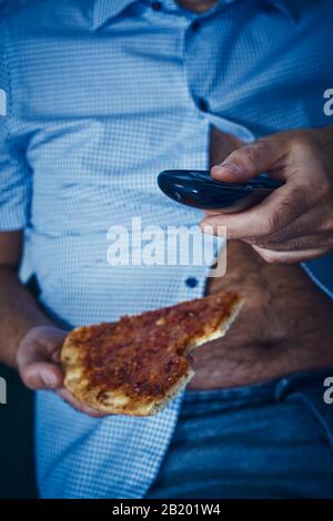 Nahaufnahme eines kaukasischen Mannes mit einem haarigen Bierbauch, auf dem Sofa sitzend, Pizza essen, während er den Fernseher beobachtet Stockfoto