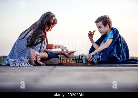 Zwei kleine, niedliche kleine Freunde, Junge und Mädchen, die Spaß haben, während sie Schach spielen, das mit einer Decke am See bedeckt ist. Kinder spielen. Freundschaft. Stockfoto