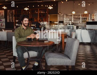Fröhlicher kaukasischer junger Mann, der mit dem Handy im Café sitzt