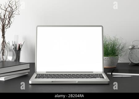 Arbeitsbereich mit leerem Laptop-Monitor mit weißem Bildschirm auf dem dunklen Schreibtisch in der Nähe der weißen Wand Stockfoto