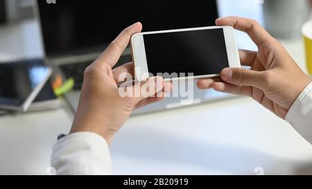 Foto der Hand des jungen Geschäftsmannes mit weißem Smartphone mit schwarzem leeren Bildschirm am modernen Arbeitstisch mit verschwommenem Laptop und Bürogeräten wie Stockfoto