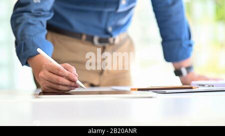 Kreativer Mann, der als Architekt am Computertablett steht und schreibt/zeichnet, der einen modernen Arbeitstisch mit komfortablem Bürobackgro-Design aufstellt Stockfoto