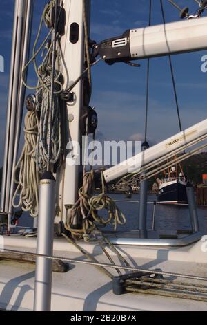 Ein Nahblick auf einen Mast, Segel und Takelage auf einem privaten Segelboot mit Fischerboot und Hafen im Hintergrund Stockfoto