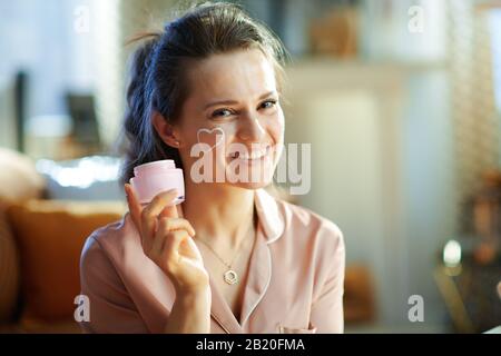 Porträt einer fröhlichen eleganten Frau im mittleren Alter in Pyjamas mit weißem Gesichtscreme Herz auf Wange zeigt geöffnetes kosmetisches Glas zu Hause in sonniger Winte Stockfoto