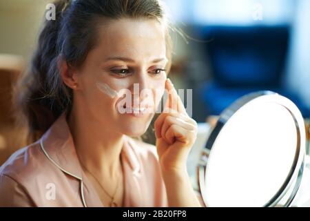 Lächelnde elegante Frau im Schlafanzug im modernen Haus am sonnigen Wintertag Gesichtscreme auftragen und im Spiegel schauen. Stockfoto