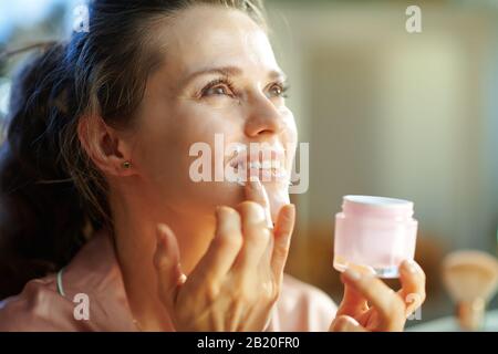 Lächelnde, stilvolle Hausfrau im mittleren Alter im Schlafanzug mit rosafarbenem Kosmetikbecher, Lippenkonturcreme im modernen Wohnzimmer am sonnigen Wintertag. Stockfoto