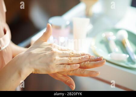 Nahaufnahme der Frau, die im sonnigen Wintertag Handcreme in der Nähe des Tisches mit Toilettenartikeln zu Hause aufbringen kann. Stockfoto