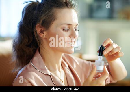 Fröhliche moderne Frau im Schlafanzug im modernen Zuhause am sonnigen Wintertag mit kosmetischem Elixier. Stockfoto