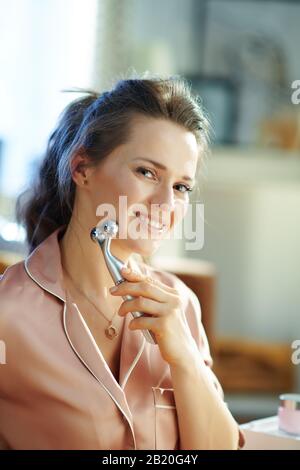 Lächelnde, stylische 40 Jahre alte Frau im Schlafanzug im modernen Wohnzimmer am sonnigen Wintertag mit einem Anti-Aging-Elektro-Massagegerät für den Gesichtsaufzug. Stockfoto