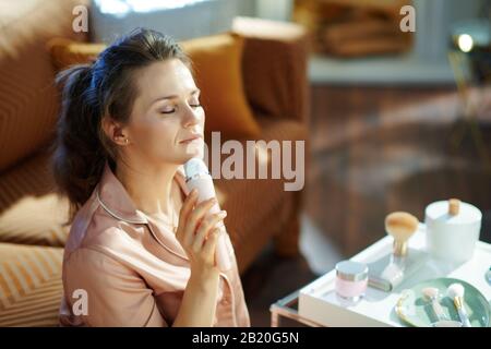 Entspannte, moderne Mittelaltersfrau im Schlafanzug mit elektrischem Gesichtsreinigungstisch, bürstennaher Tisch mit Toilettenartikeln im modernen Wohnzimmer im sonnigen Winter d Stockfoto