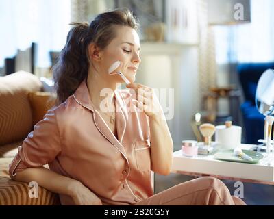 Entspannte, moderne 40 Jahre alte Frau im Schlafanzug mit beruhigendem Quarzrollen-Massagegerät in der Nähe eines Tisches mit Toilettenartikeln im modernen Wohnzimmer im sonnigen Winter Stockfoto