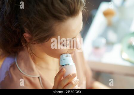 Elegantes Weibchen im Schlafanzug im modernen Zuhause am sonnigen Wintertag mit elektrischer Gesichtsreinigungsbürste. Stockfoto