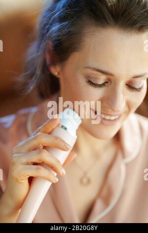 Lächelnde, stilvolle Hausfrau im mittleren Alter im Schlafanzug im modernen Wohnzimmer am sonnigen Wintertag mit elektrischer Gesichtsreinigungsbürste. Stockfoto