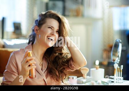 Lächelnde, stylische Frau im Schlafanzug mit langem, welligem Haar, das eine Haarölflasche in der Nähe eines Tisches mit Toilettenartikeln im modernen Haus im sonnigen Winter hält d Stockfoto