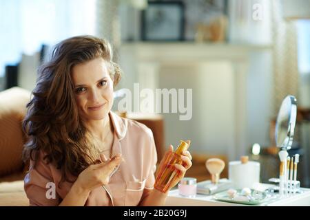 Porträt der eleganten 40 Jahre alten Frau im Schlafanzug mit langem, welligem Haar, das die Haarölflasche hält und Haarspitzen in der Nähe des Tisches mit Toilettenartikeln überprüft Stockfoto