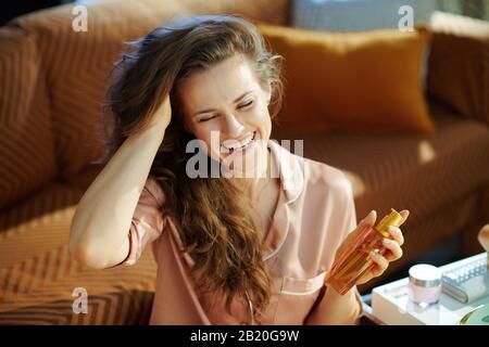 Lächelnde, stilvolle Hausfrau im Schlafanzug mit langen, welligen, brünetten Haaren, die im modernen Haus am sonnigen Wintertag die Haarölflasche halten. Stockfoto