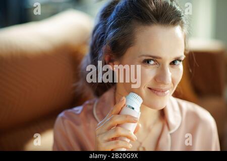 Lächelnde junge Hausfrau im Schlafanzug im modernen Zuhause am sonnigen Wintertag mit elektrischer Gesichtsreinigungsbürste. Stockfoto