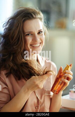Porträt einer fröhlichen, stylischen Frau im Schlafanzug mit langen, welligen Haaren, die die Haarölflasche halten und Haarspitzen im sonnigen Winter im modernen Zuhause prüfen Stockfoto