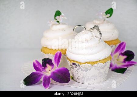 Hochzeitsringe in weißer Aufschäumung auf Cupcake mit Orchideenblüten Stockfoto