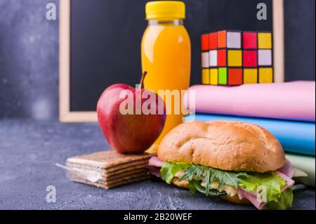 Mittagessen in der Schule in einer Schachtel in der Nähe der Tafel an der Schule. Sandwich mit Salat und Käse, eine Flasche Saft, ein apfel, Bücher, ein Wecker. Gesunde Nahrung für den Schuljungen. Freier Speicherplatz für Text Stockfoto