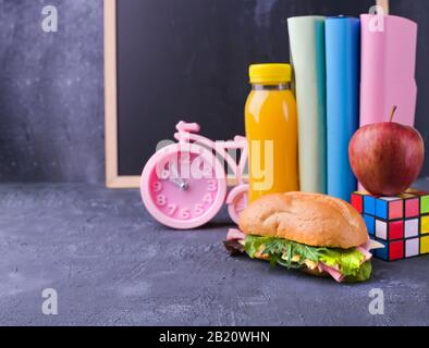 Mittagessen in der Schule in einer Schachtel in der Nähe der Tafel an der Schule. Sandwich mit Salat und Käse, eine Flasche Saft, ein apfel, Bücher, ein Wecker. Gesunde Nahrung für den Schuljungen. Freier Speicherplatz für Text Stockfoto