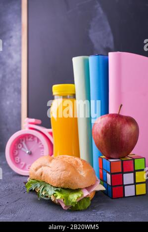 Mittagessen in der Schule in einer Schachtel in der Nähe der Tafel an der Schule. Sandwich mit Salat und Käse, eine Flasche Saft, ein apfel, Bücher, ein Wecker. Gesunde Nahrung für den Schuljungen. Freier Speicherplatz für Text Stockfoto