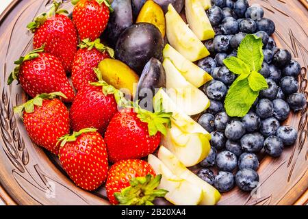 Rohe Früchte Beeren Platte, apfel, Erdbeere, Blaubeere, Pflaume, Minze. Auf ethnischer Keramikplatte, gesundes vegetarisches (veganes) Lebensmittelkonzept. Steinbeton Stockfoto