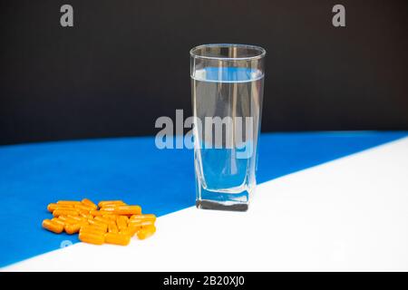Ein Glas Wasser steht neben mehreren orangefarbenen Pillen auf weiß-blauem Grund Stockfoto