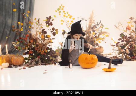 Kleines süßes Mädchen mit langen Haaren, das in schwarzem Kleid und Hut mit herbstlichen Dekorationen im Hintergrund während Halloween auf Szene steht Stockfoto