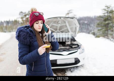 Junge Frauen, die eine Mitgliedschaft in der Straßenhilfe oder eine Visitenkarte besitzen und neben einem kaputten Auto mit geöffneter Haube Hilfe anfordern. Wintersaison. Stockfoto