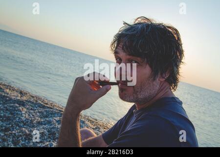 Charmanter Mann mittleren Alters mit einer Zigarrenbestuhlung am Strand bei Sonnenuntergängen/Nahporträts Stockfoto
