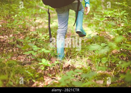 Schneiden Sie Beine von weiblichen Reisenden in Jeans und Gummistiefeln mit Rucksack auf Gelände mit hohem grünen Gras in Wald Stockfoto