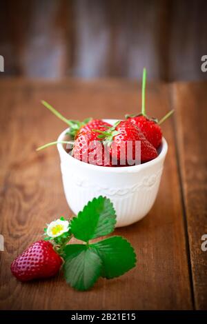 Reife rote Erdbeeren mit grünen Blättern in weißer Schüssel auf Holztisch Stockfoto