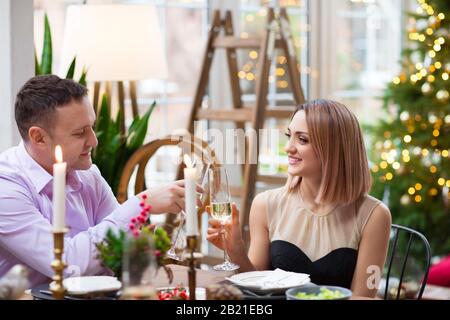 Fröhliches junges Paar in elegantem Anzug und Kleid sitzt am festlichen Tisch mit Gläsern in den Händen lächelnd und sieht sich im modernen Wohnzimmer an Stockfoto