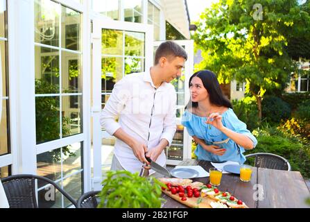 Junger Mann und Frau lächeln und reden miteinander, während sie am Sommertag im Garten Bruschetten auf den Tisch bereiten Stockfoto