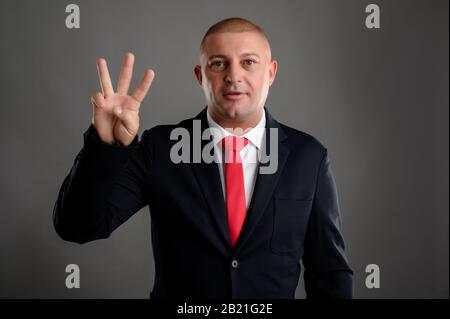 Porträt eines coolen Geschäftsmannes mit elegantem schwarzen Anzug und roter Krawatte, die drei isoliert über grauem Hintergrund zeigt Stockfoto